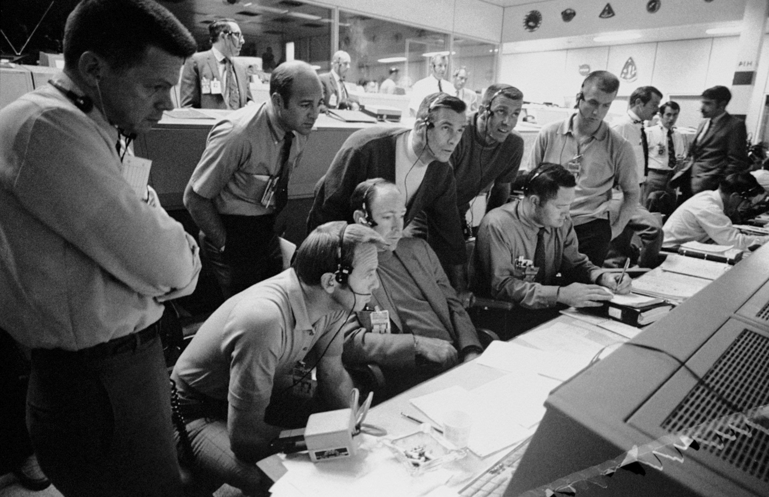 In diesem am 14. April 1970 aufgenommenen Foto von NASA, beobachtet eine Gruppe von sechs Astronauten und zwei Flugleitern die Konsolenaktivität im Mission Operations Control Room (MOCR) des Mission Control Center (MCC) während der problembehafteten Apollo 13 Mission zum Mond. 
