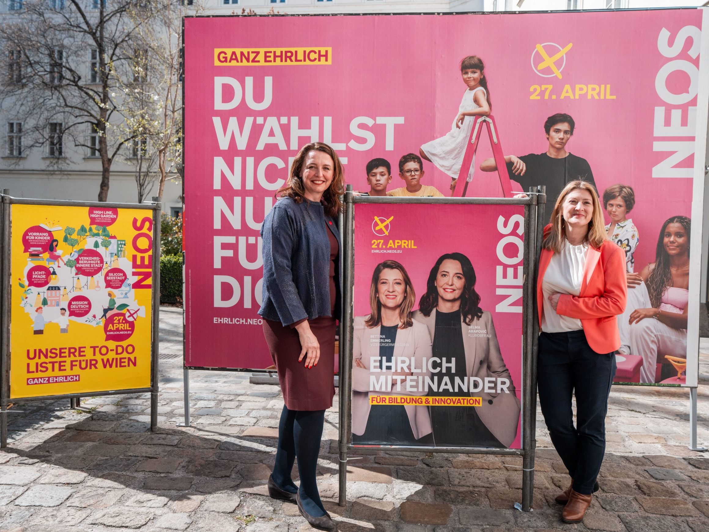 Die NEOS-Spitzenkandidatinnen für die Wien-Wahl neben einem Plakat mit ihnen. Die NEOS-Spitzenkandidatinnen für die Wien-Wahl neben einem Plakat mit ihnen.