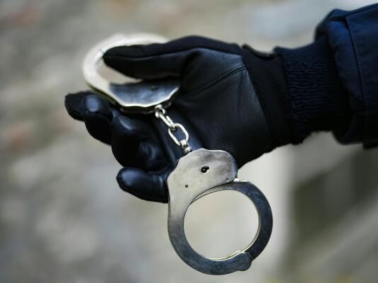 Auf dem Foto sieht man Handschellen. Sie werden von einer Hand mit Handschuhen gehalten.