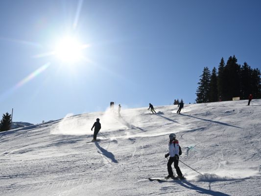 Auf dem Bild sieht man einige Skifahrer. Sie fahren eine Piste hinunter. Es scheint die Sonne und der Himmel ist blau.